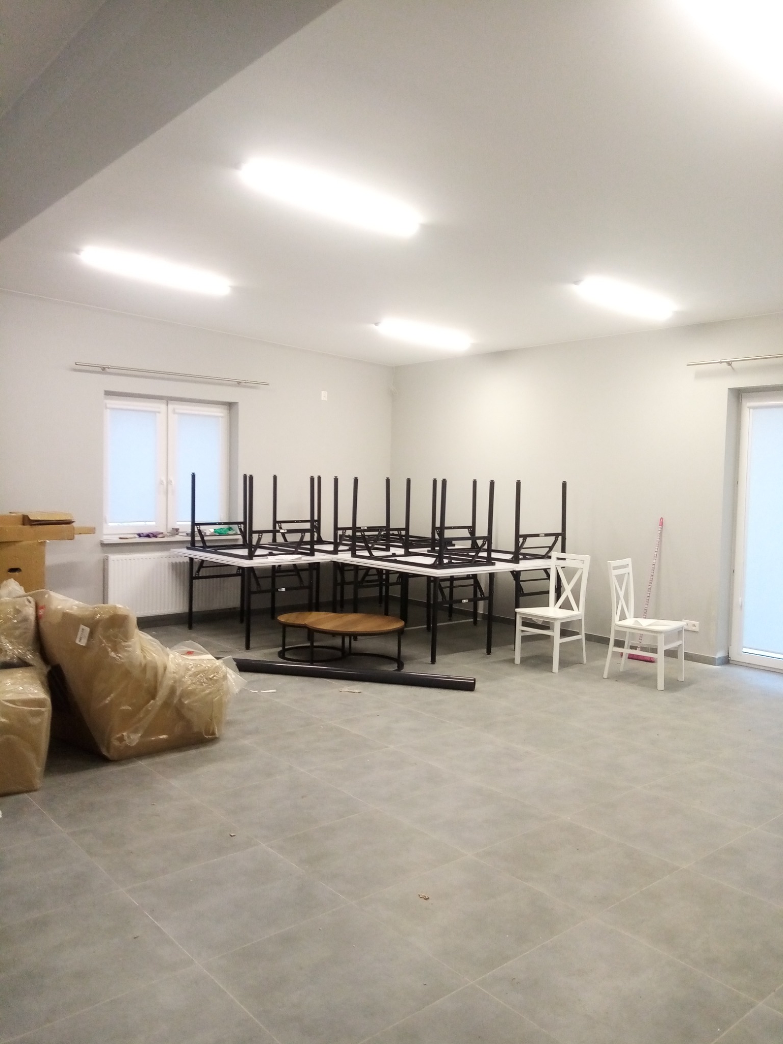 Puste pomieszczenie z szarymi płytkami podłogowymi, białymi ścianami, oświetleniem sufitowym, oknem z roletą, oraz meblami w trakcie montażu lub demontażu: odwrócone stoły na stołach, białe krzesła...