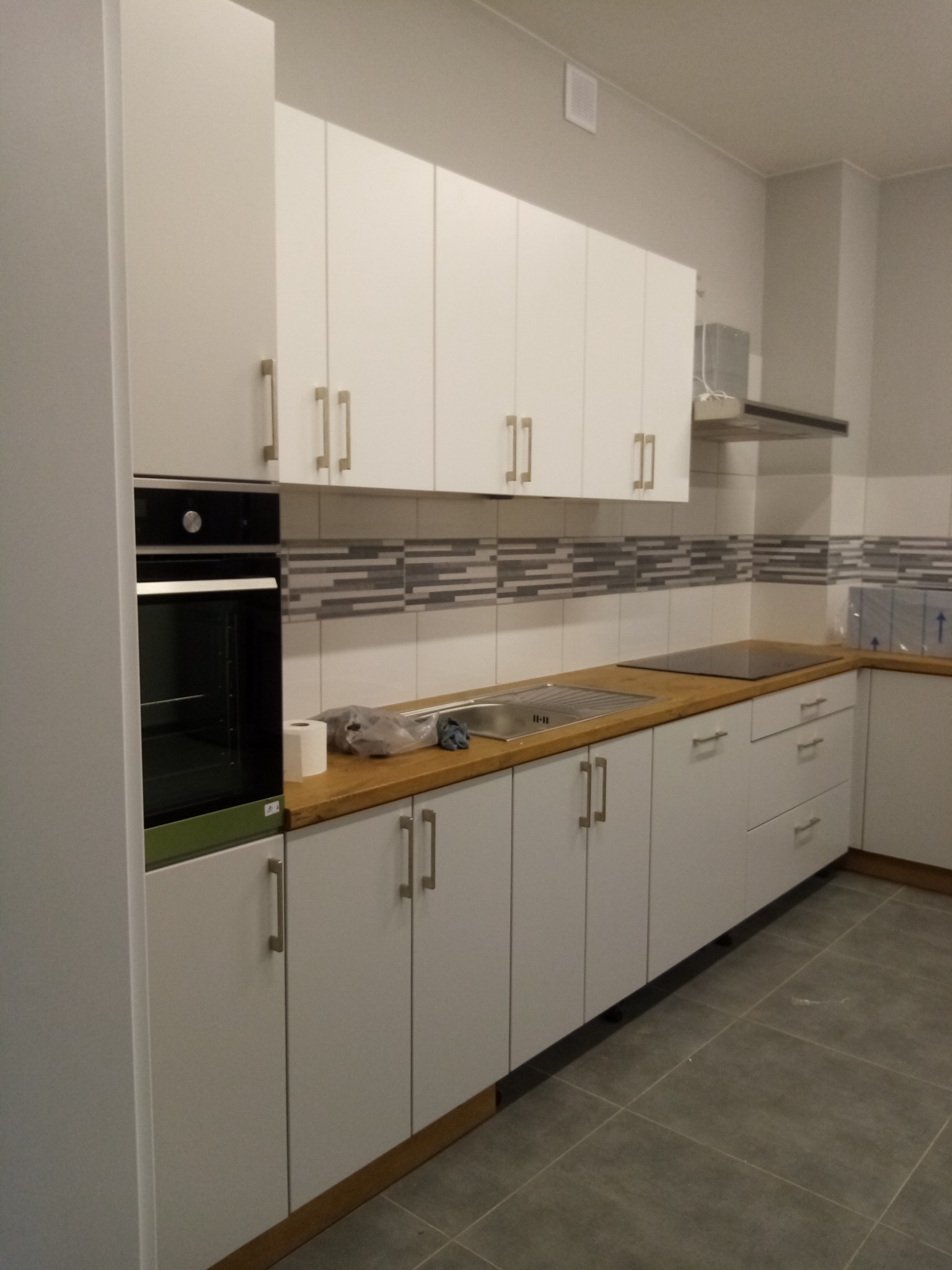 Nowoczesna kuchnia z białymi szafkami, drewnianym blatem i szarą mozaiką na ścianie, widok na zabudowany piekarnik i płytę grzewczą.