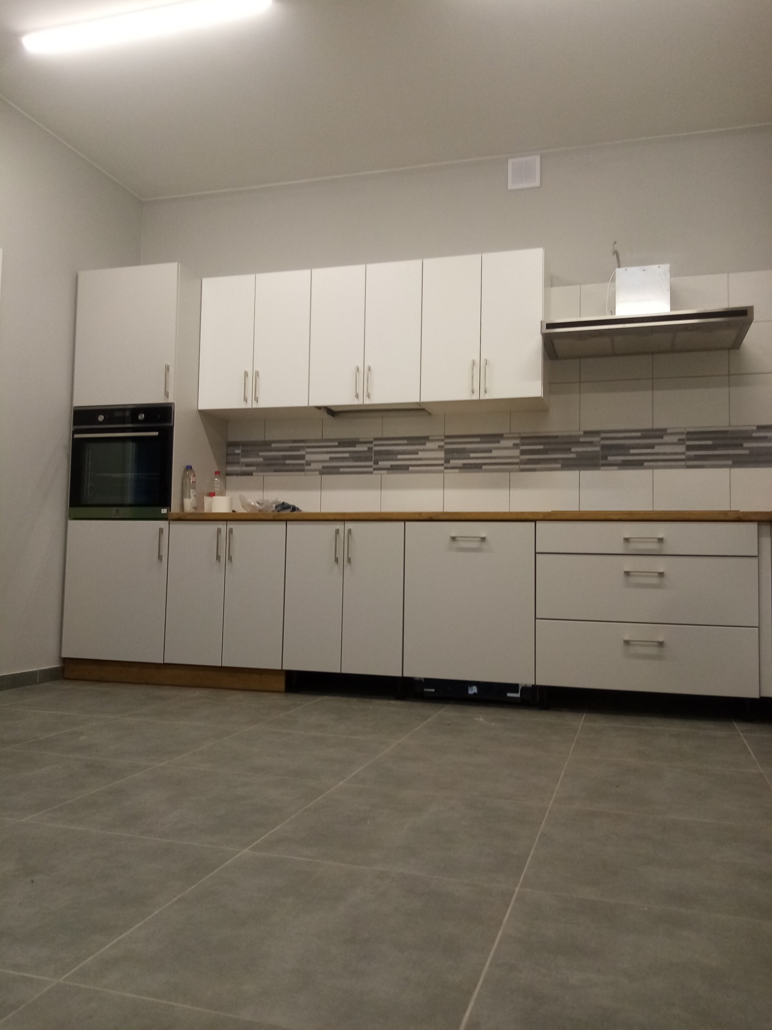 Jasna kuchnia z białymi szafkami i szarymi płytkami na ścianie, widok z niskiego kąta, szara podłoga z płytek.