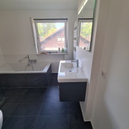 KUBA BAU - Nowoczesna łazienka na poddaszu z czarną płytką podłogową, białą wanną obudowaną czarnymi płytkami, umywalką z szafką i lustrem. Okno dachowe z widokiem na drzewa i dom po drugiej stronie ulicy.