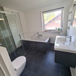 KUBA BAU - Nowoczesna łazienka z czarną podłogą, białą wanną obudowaną szarą płytką, przeszklonym prysznicem i umywalką z szafką, widok z perspektywy drzwi.