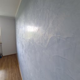 KUBA BAU - Ściana z efektem dekoracyjnym w odcieniach szarości i bieli, widoczne refleksy światła, drewniana podłoga i fragment okna.