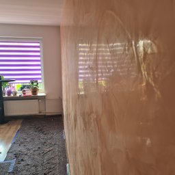 KUBA BAU - Ściana z efektem dekoracyjnym w odcieniach brązu i beżu, zabezpieczona podłoga z widocznym oknem z żaluzjami w tle.