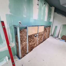 KUBA BAU - Ściana w trakcie zabudowy z płyt gipsowo-kartonowych, widoczne profile metalowe, izolacja z wełny mineralnej, częściowo zagruntowane płyty, czerwona poziomica przy ścianie, instalacja elektryczna...