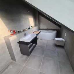 Szare płytki podłogowe i ścienne w łazience na poddaszu, biała wanna narożna i podwieszana toaleta, umywalka nablatowa na czarnej szafce, widoczne akcesoria na półce.