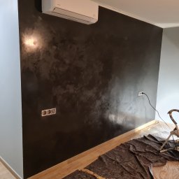 KUBA BAU - Ściana z efektem dekoracyjnym w odcieniach szarości i brązu, widoczne gniazdka elektryczne, drabina malarska i folia ochronna na podłodze, klimatyzator na górze ściany.