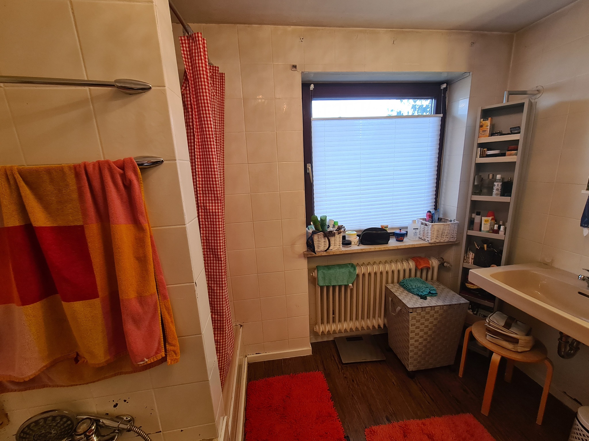Umeblowana łazienka z beżowymi płytkami, czerwonymi dywanikami i zasłoną w kratę, widoczne ślady użytkowania i drobne niedoskonałości na ścianach i fugach.