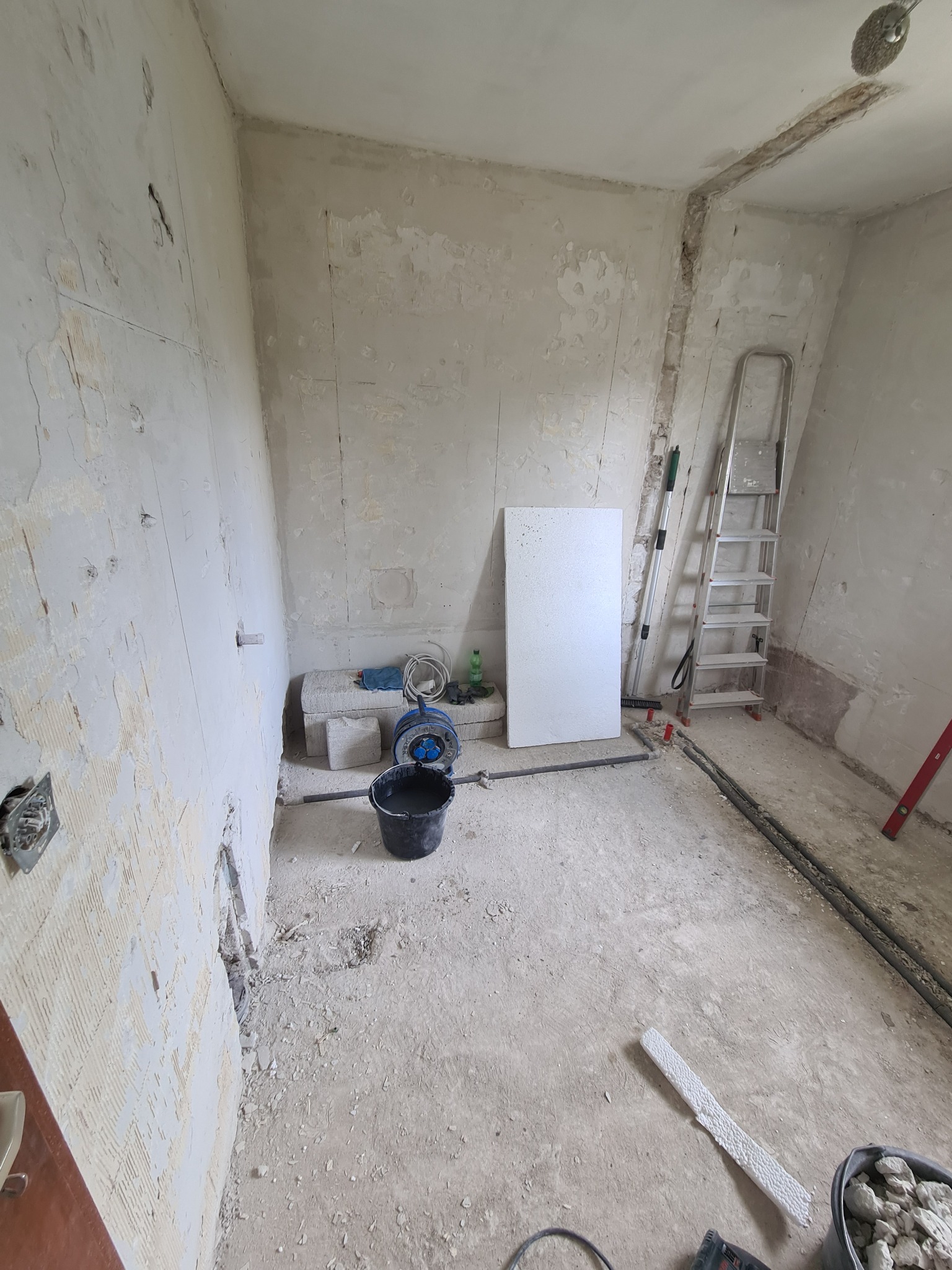 Pokój w trakcie remontu: ściany ze śladami starej farby i tynku, widoczne instalacje elektryczne, drabina, wiadro z zaprawą, agregat malarski i płyta styropianowa.