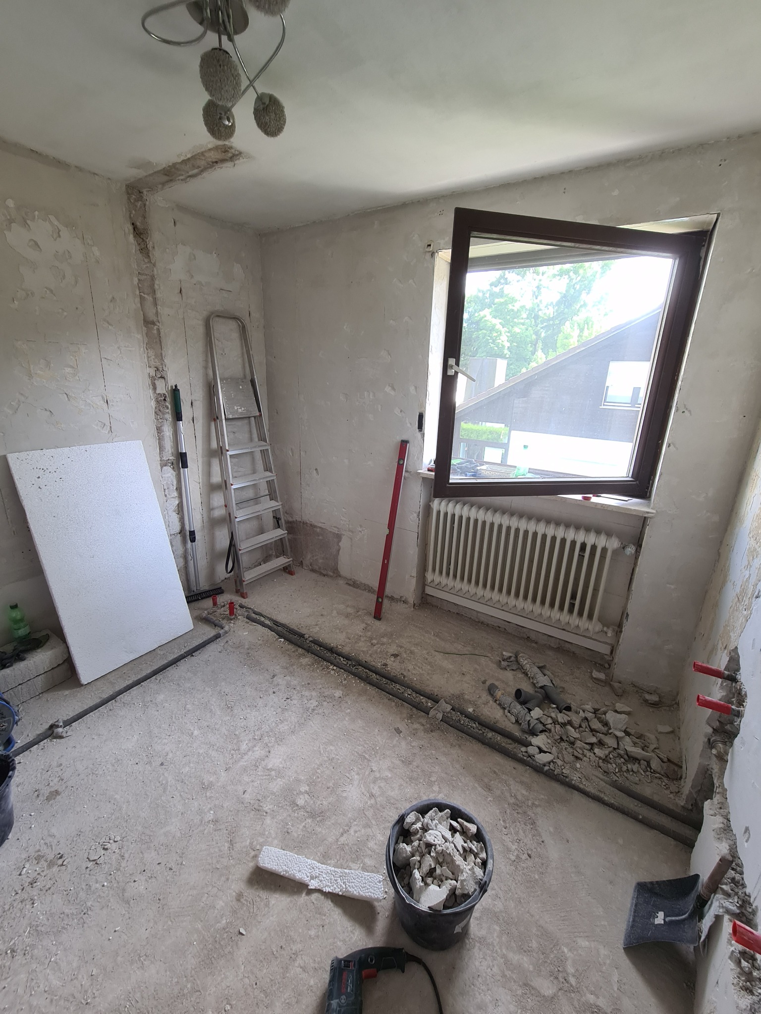 Pokój w trakcie remontu: odsłonięte ściany, rury, gruz w wiadrze, drabina oparta o ścianę, otwarte okno z widokiem na zieleń.