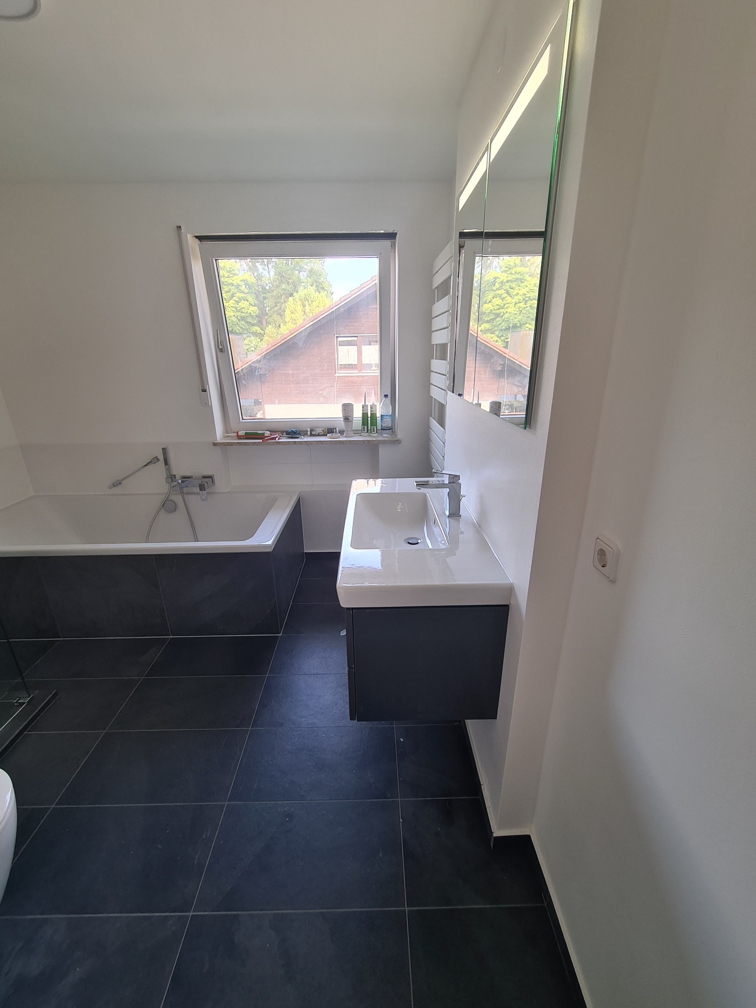Nowoczesna łazienka na poddaszu z czarną płytką podłogową, białą wanną obudowaną czarnymi płytkami, umywalką z szafką i lustrem. Okno dachowe z widokiem na drzewa i dom po drugiej stronie ulicy.
