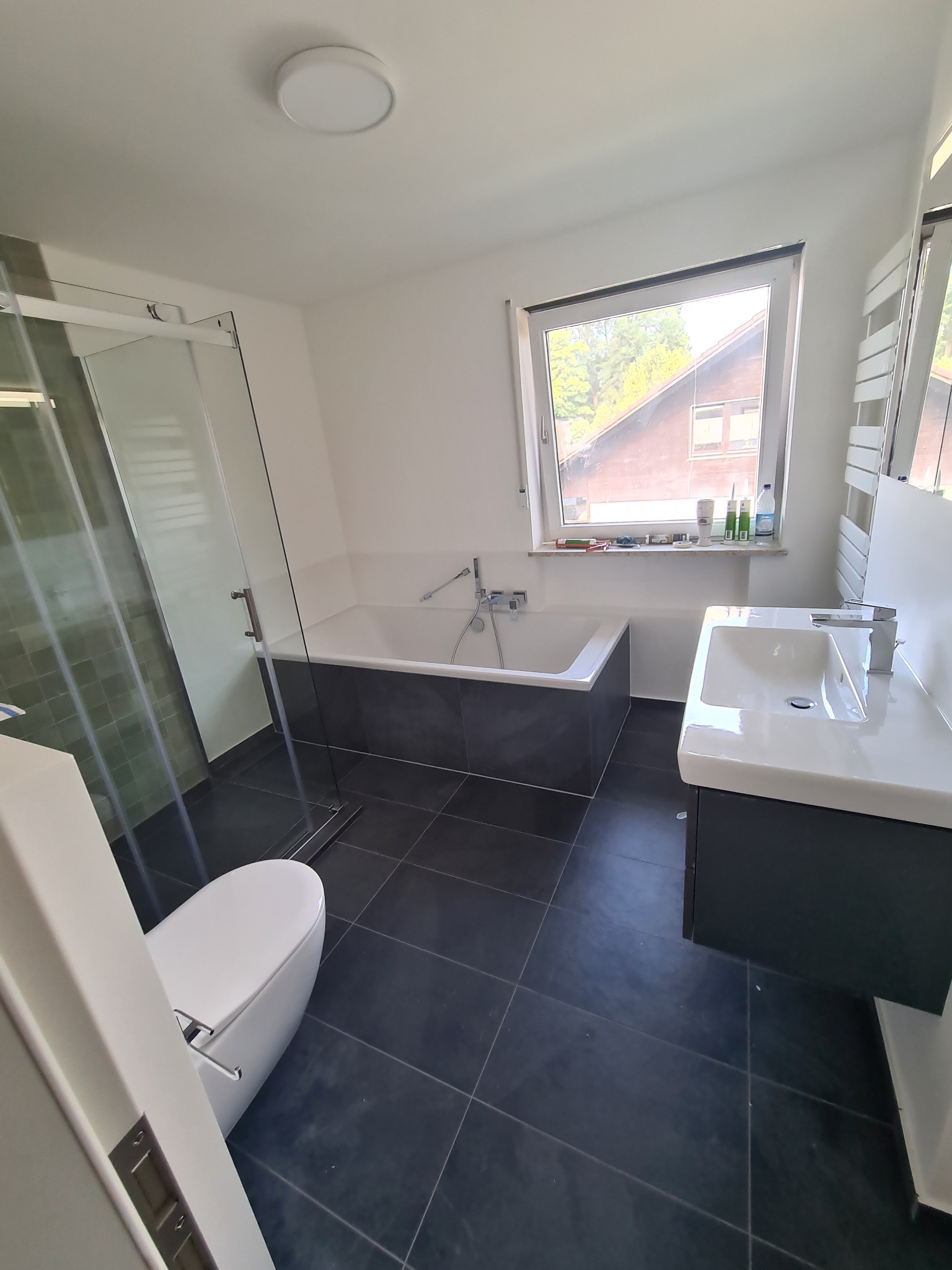 Nowoczesna łazienka z czarną podłogą, białą wanną obudowaną szarą płytką, przeszklonym prysznicem i umywalką z szafką, widok z perspektywy drzwi.