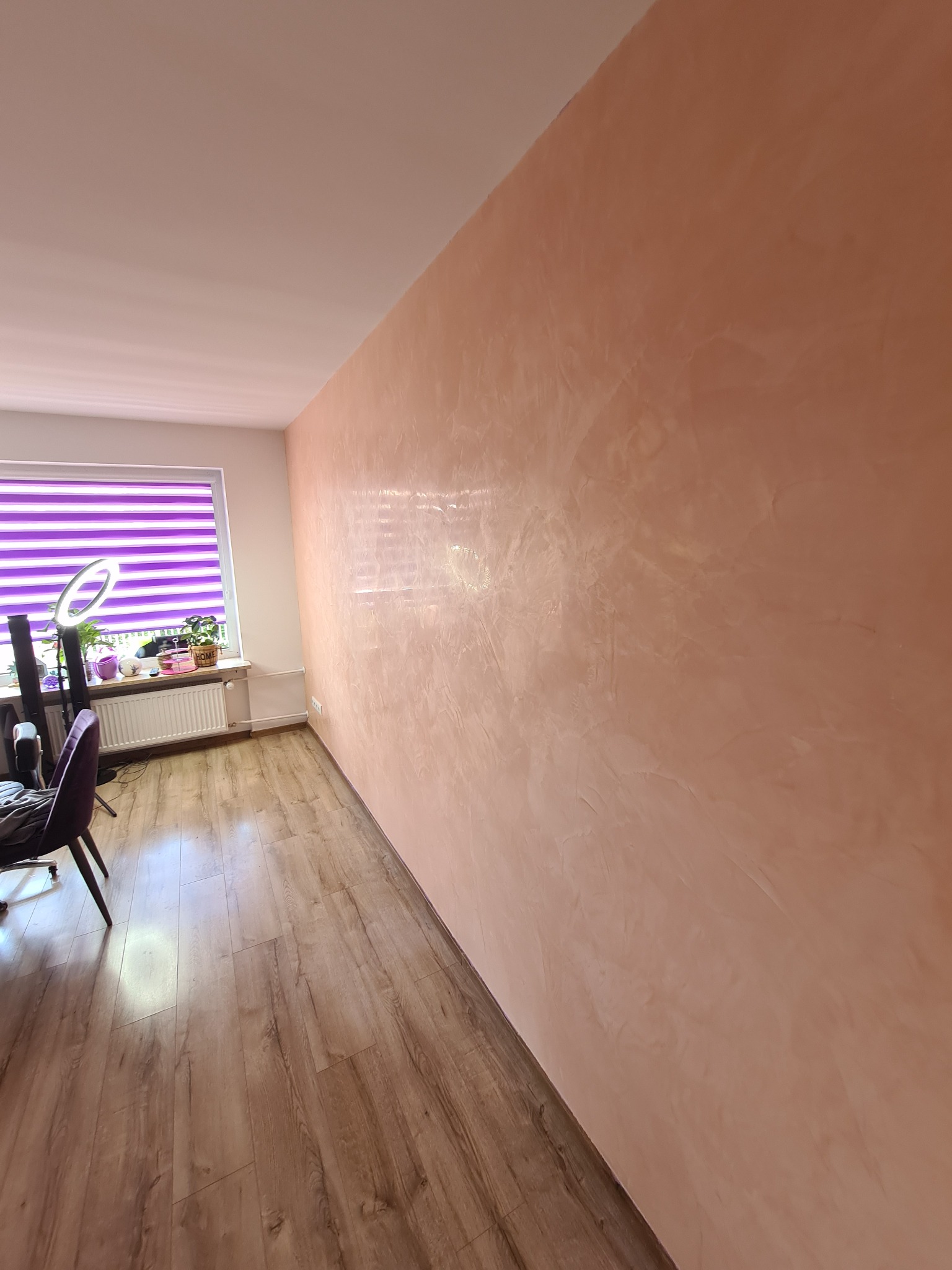 Ściana w odcieniach brzoskwini z dekoracyjną fakturą, drewnopodobna podłoga, okno z fioletowymi żaluzjami oraz biurko z lampą pierścieniową w tle.