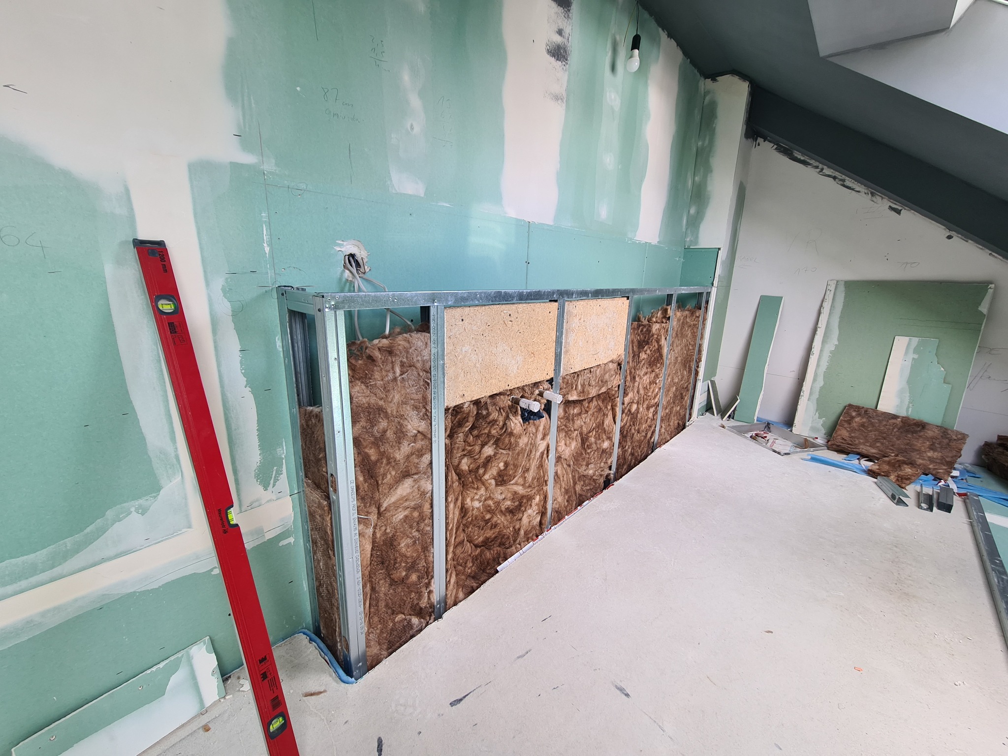 Ściana w trakcie zabudowy z płyt gipsowo-kartonowych, widoczne profile metalowe, izolacja z wełny mineralnej, częściowo zagruntowane płyty, czerwona poziomica przy ścianie, instalacja elektryczna...