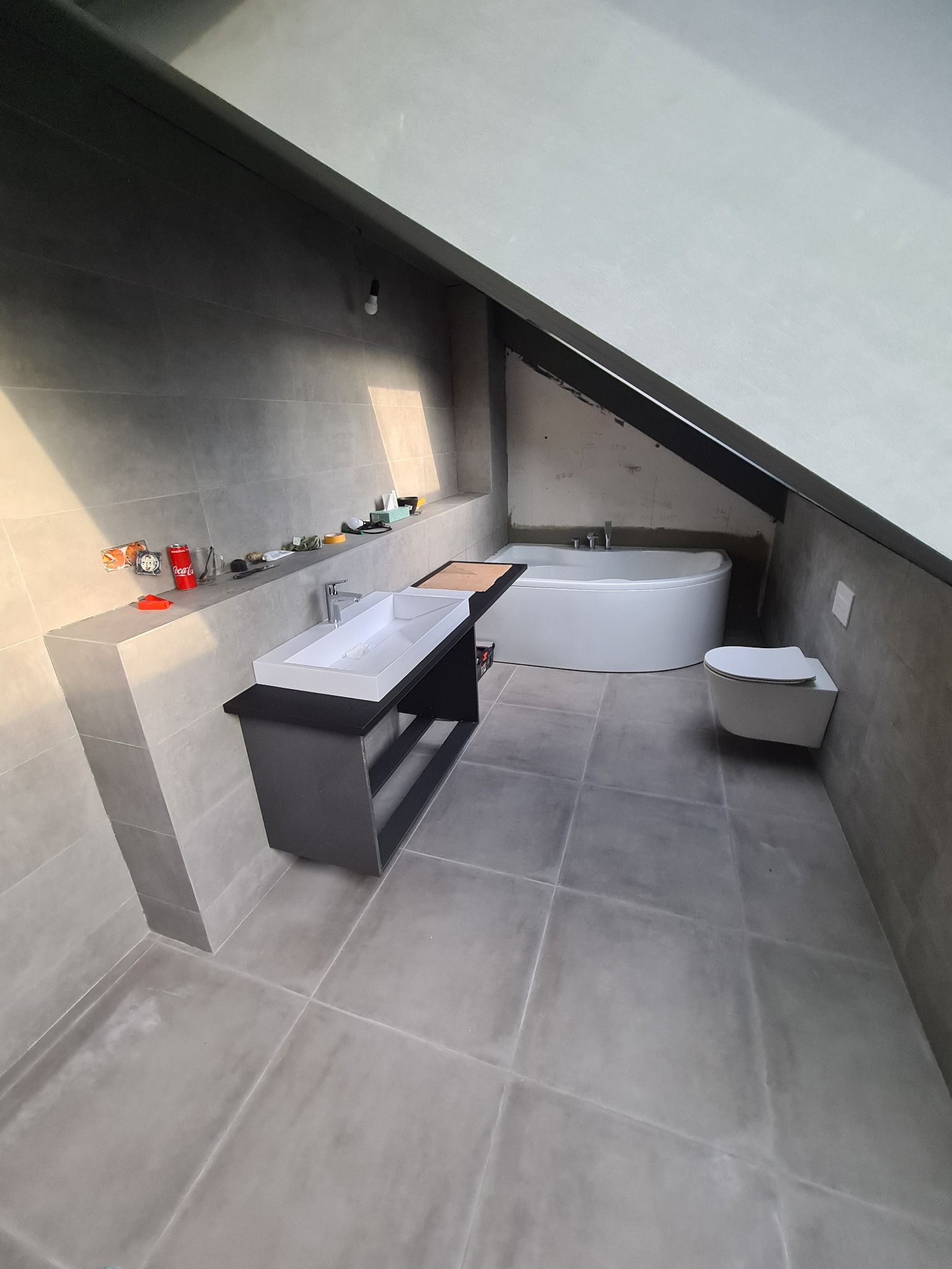 Szare płytki podłogowe i ścienne w łazience na poddaszu, biała wanna narożna i podwieszana toaleta, umywalka nablatowa na czarnej szafce, widoczne akcesoria na półce.