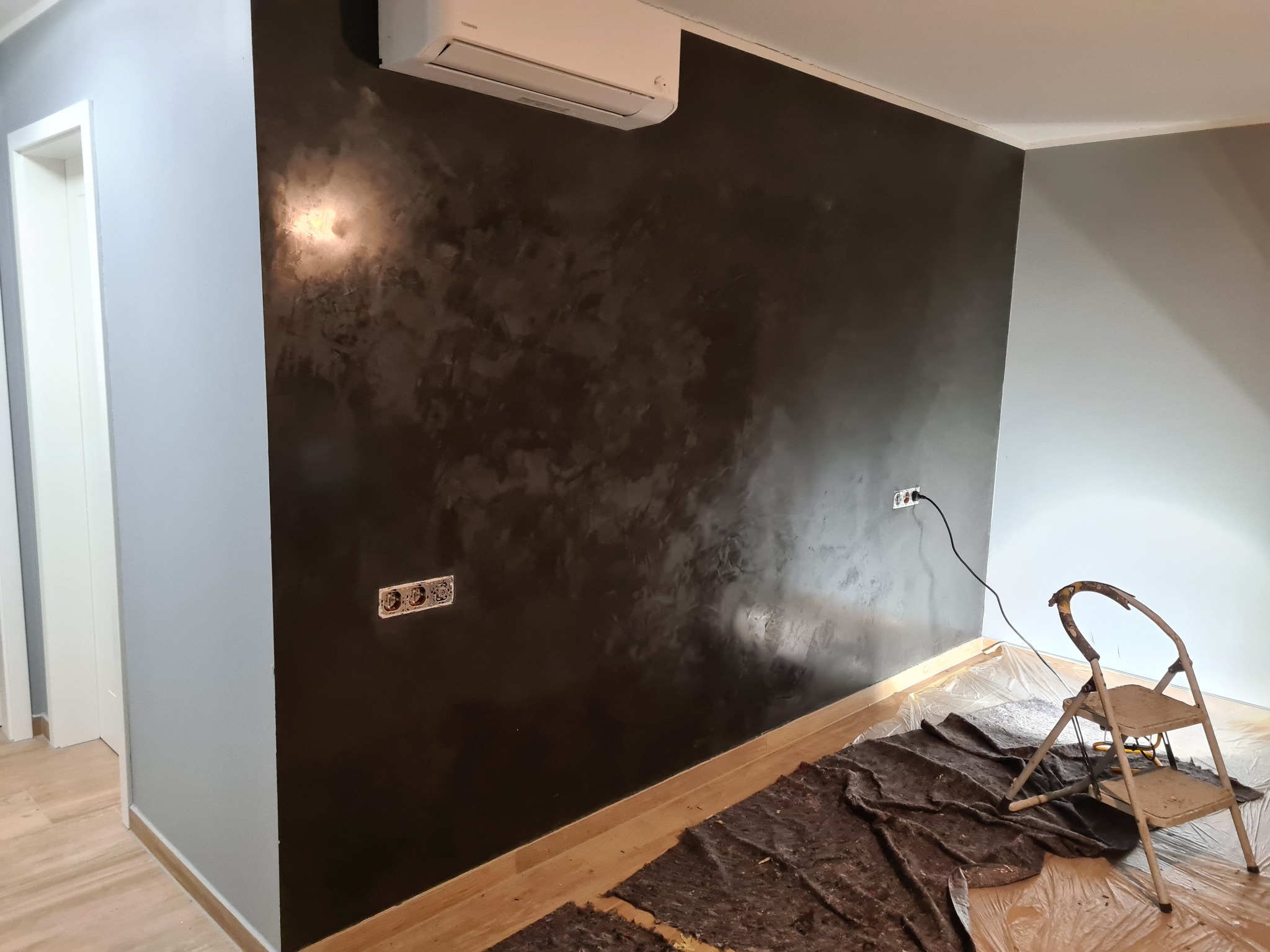 Ściana z efektem dekoracyjnym w odcieniach szarości i brązu, widoczne gniazdka elektryczne, drabina malarska i folia ochronna na podłodze, klimatyzator na górze ściany.