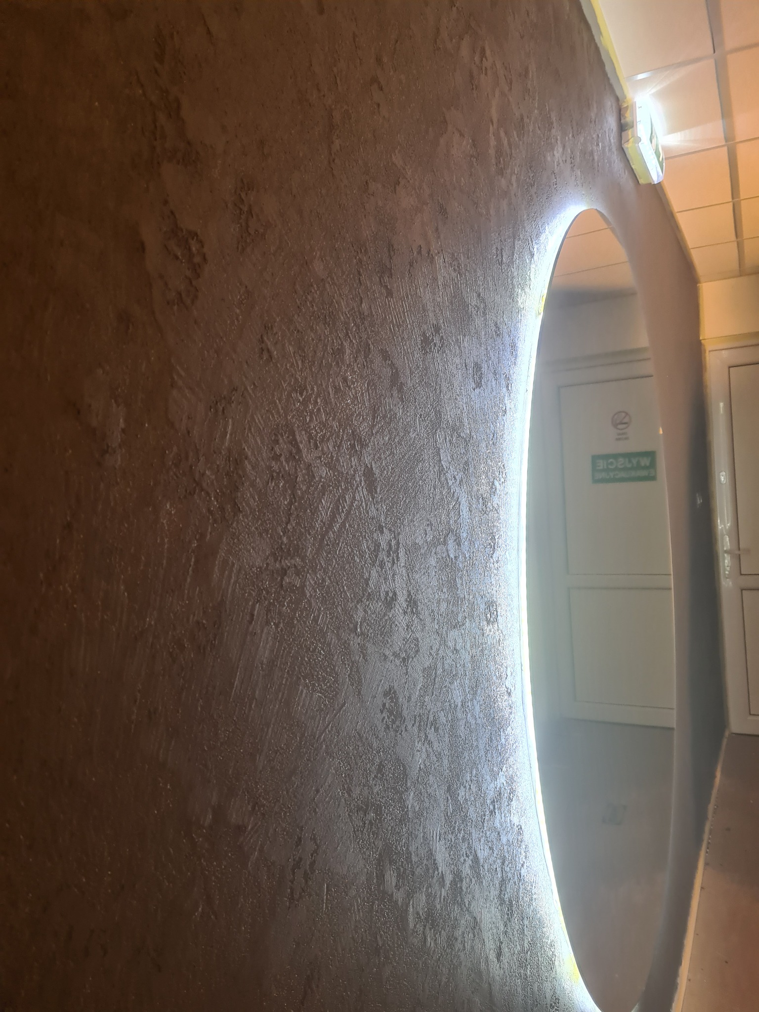 Brązowa ściana ze strukturą, oświetlona obok owalnego otworu w ścianie, widok na korytarz z białą ścianą i drzwiami.