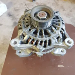 Używany alternator samochodowy z widocznymi śladami eksploatacji, umieszczony na brązowym blacie, obok widoczne narzędzia.