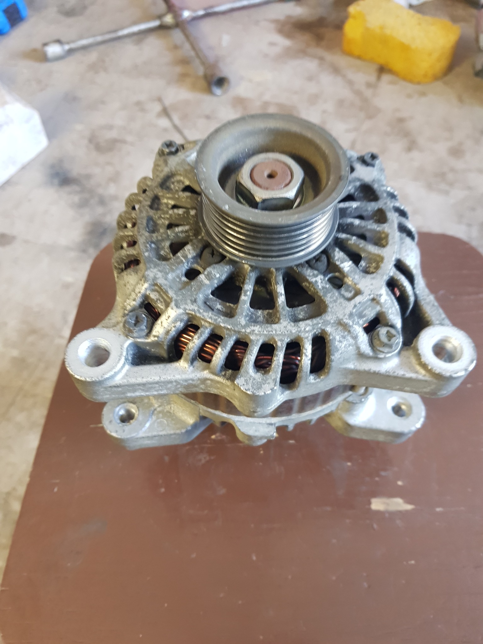 Używany alternator samochodowy z widocznymi śladami eksploatacji, umieszczony na brązowym blacie, obok widoczne narzędzia.