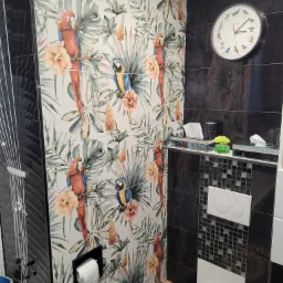 Wnętrze łazienki z czarnymi płytkami i motywem tropikalnym na ścianie, wzór z papugami i liśćmi, wiszący zegar z wizerunkami ptaków, czarne dodatki i mozaika przy spłuczce.