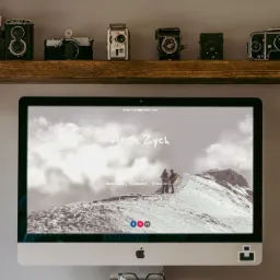 Ekran komputera iMac wyświetlający portfolio internetowe developera, filmowca i podróżnika, z adresem e-mail i zdjęciem górskim w tle, ustawiony na drewnianym stojaku z okularami i klawiaturą,...