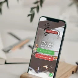 Smartfon w drewnianym uchwycie prezentuje stronę internetową China Bar Czarnk&oacute;w z logiem smoka i przyciskami do kontaktu i menu, na tle rozmytej zieleni i notatnika.