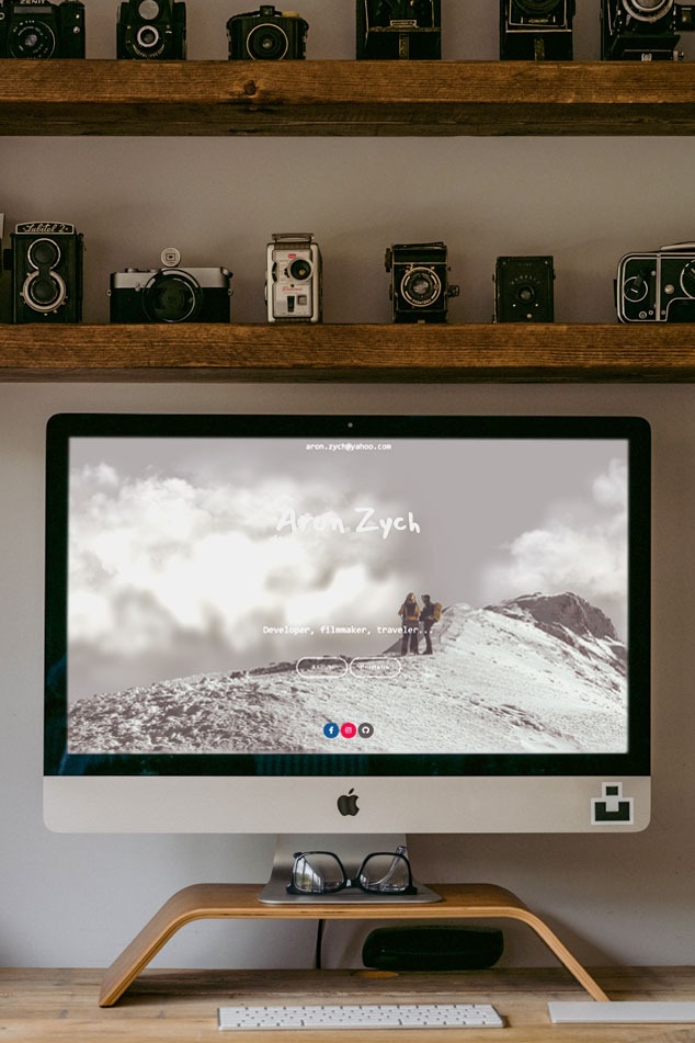 Ekran komputera iMac wyświetlający portfolio internetowe developera, filmowca i podróżnika, z adresem e-mail i zdjęciem górskim w tle, ustawiony na drewnianym stojaku z okularami i klawiaturą,...