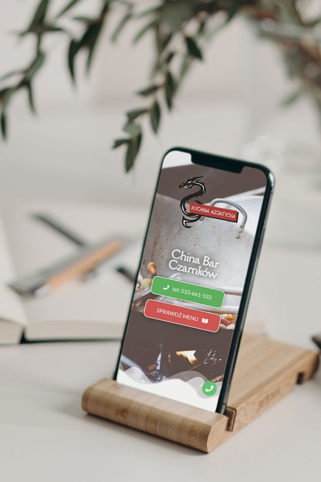 Smartfon w drewnianym uchwycie prezentuje stronę internetową China Bar Czarnków z logiem smoka i przyciskami do kontaktu i menu, na tle rozmytej zieleni i notatnika.