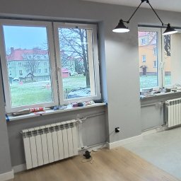 Przemysław Borysiak - Twoj Nowy Dom usługi remontowo budowlane - Szpachlowane ściany, grzejniki pod oknami i fragment ułożonej podłogi w pokoju. Na ścianie częściowo położone płytki. Widok z okna na zewnątrz.