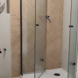 Przemysław Borysiak - Twoj Nowy Dom usługi remontowo budowlane - Nowoczesna kabina prysznicowa z czarnymi ramami na białym brodziku, wykończona płytkami w beżowe pasy. Widoczne przyłącza wodne.