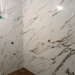 Białe płytki imitujące marmur z wyraźnym żyłowaniem w odcieniach szarości i złota, na ścianie widoczne otwory instalacyjne, na podłodze panele imitujące drewno.