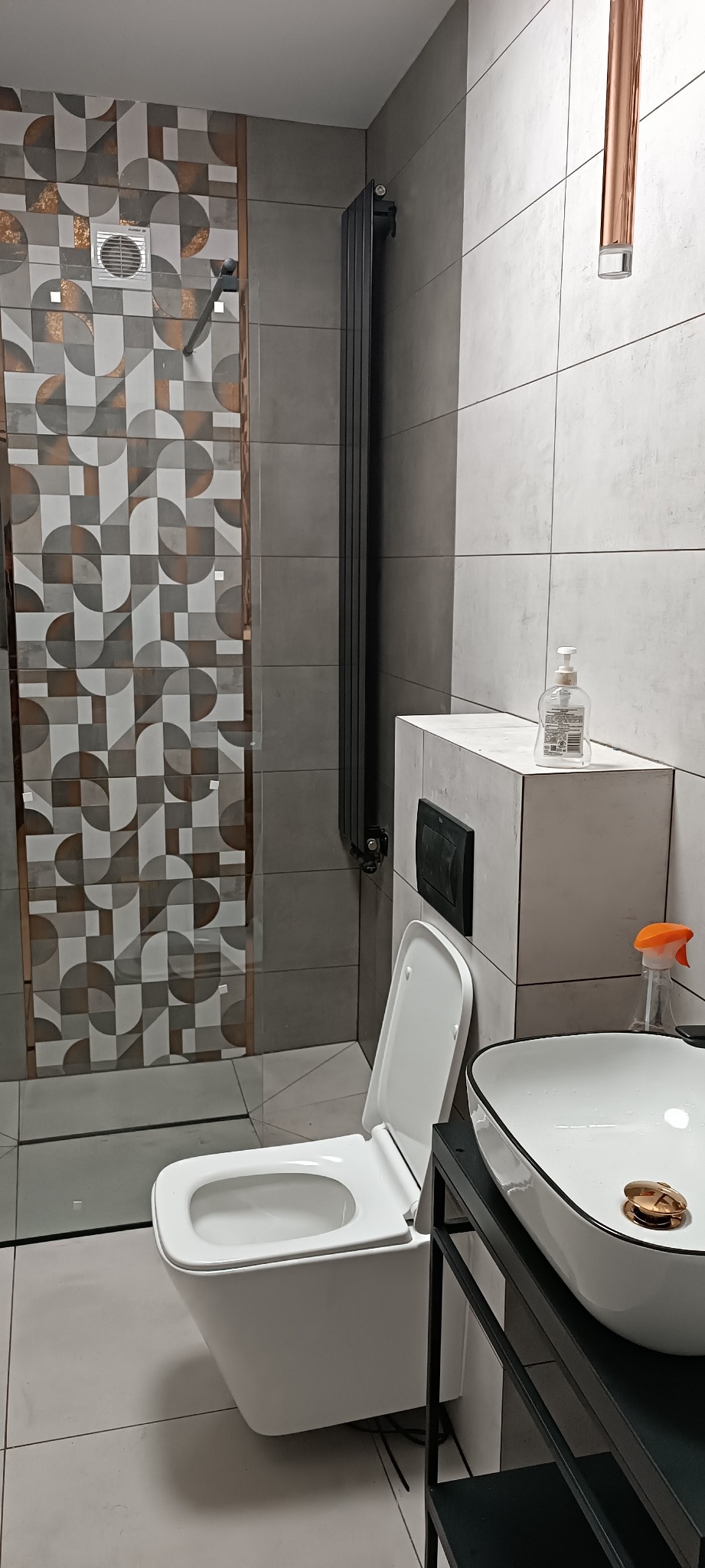 Nowoczesna łazienka z wiszącą toaletą, szklaną kabiną prysznicową z wzorzystymi płytkami i grzejnikiem drabinkowym. Umywalka nablatowa z czarnym stelażem.