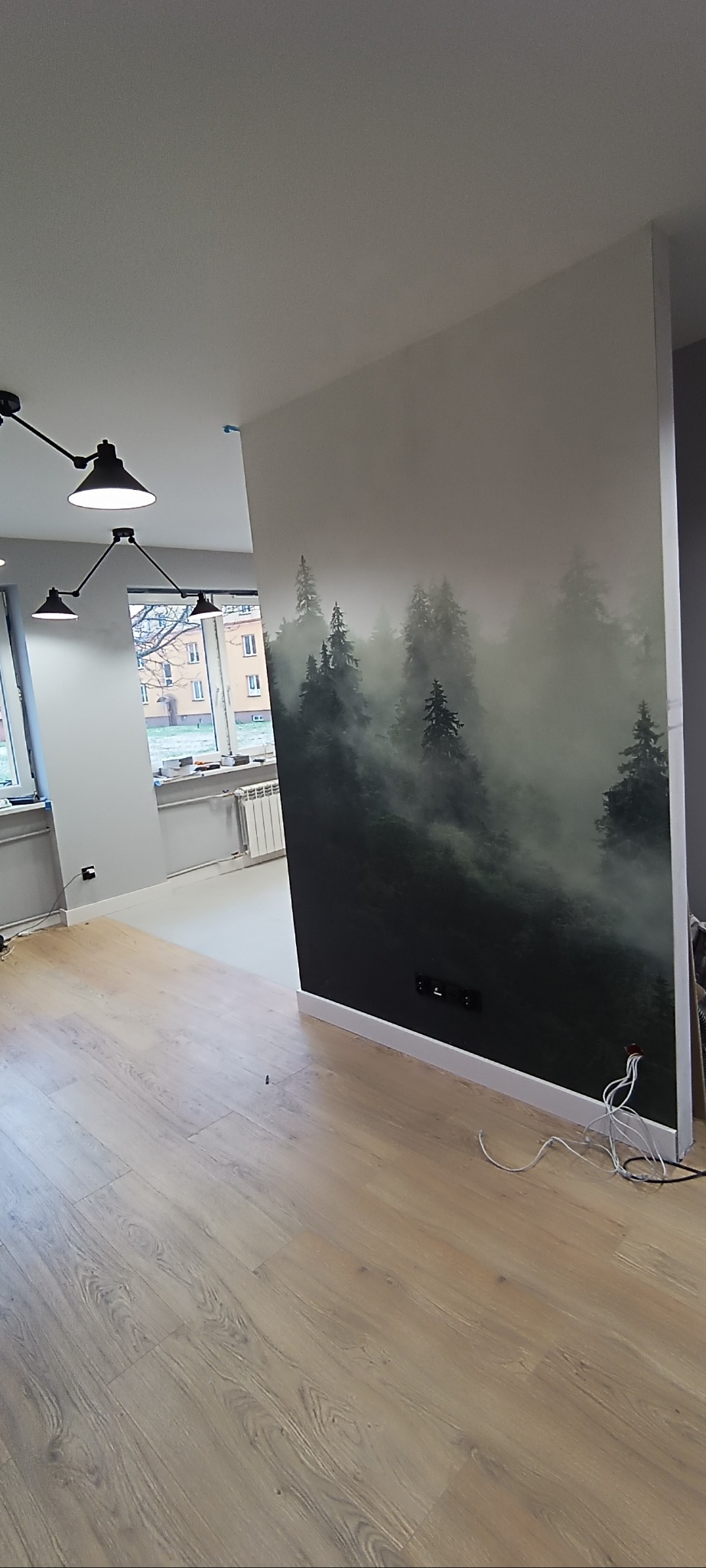 Wnętrze po remoncie: jasne panele, ściana z fototapetą lasu we mgle, designerska lampa sufitowa. Widok na sąsiednie pomieszczenie z oknem.