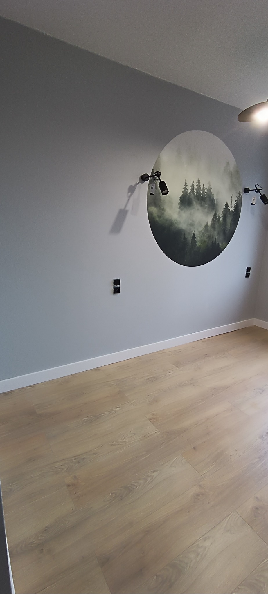 Jasne panele podłogowe w pokoju z szarą ścianą, na której widnieje okrągła grafika lasu we mgle i czarne reflektory. Widoczne listwy przypodłogowe i gniazdka.