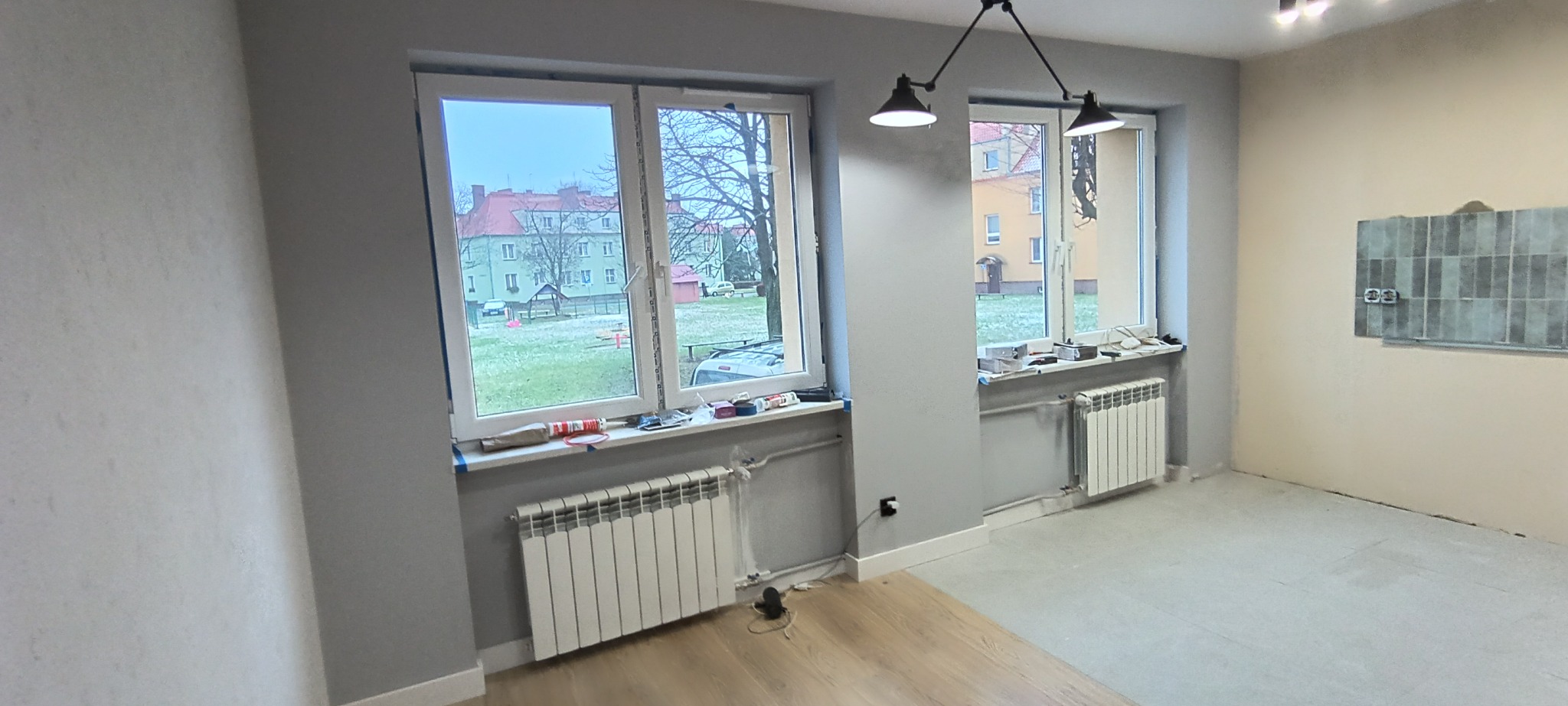 Szpachlowane ściany, grzejniki pod oknami i fragment ułożonej podłogi w pokoju. Na ścianie częściowo położone płytki. Widok z okna na zewnątrz.