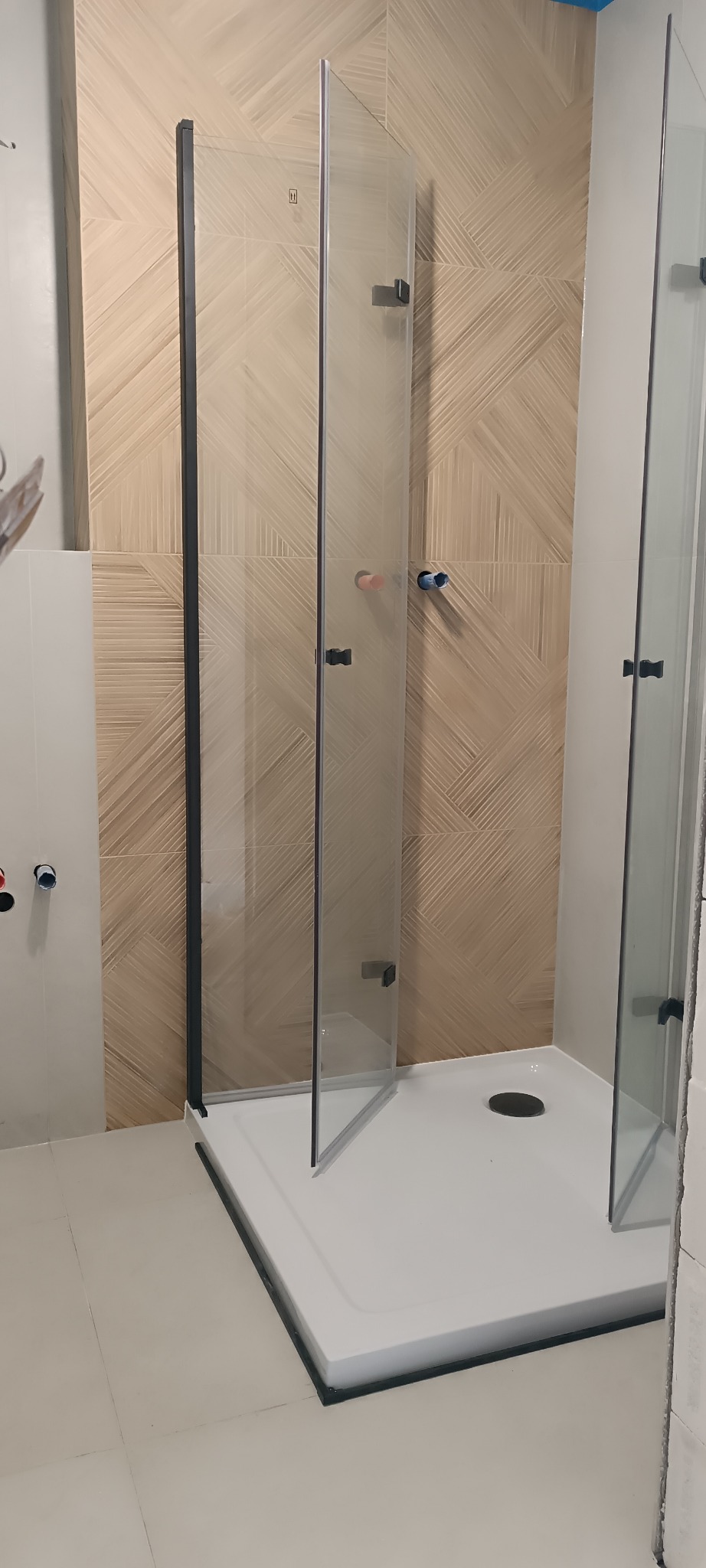 Nowoczesna kabina prysznicowa z czarnymi ramami na białym brodziku, wykończona płytkami w beżowe pasy. Widoczne przyłącza wodne.