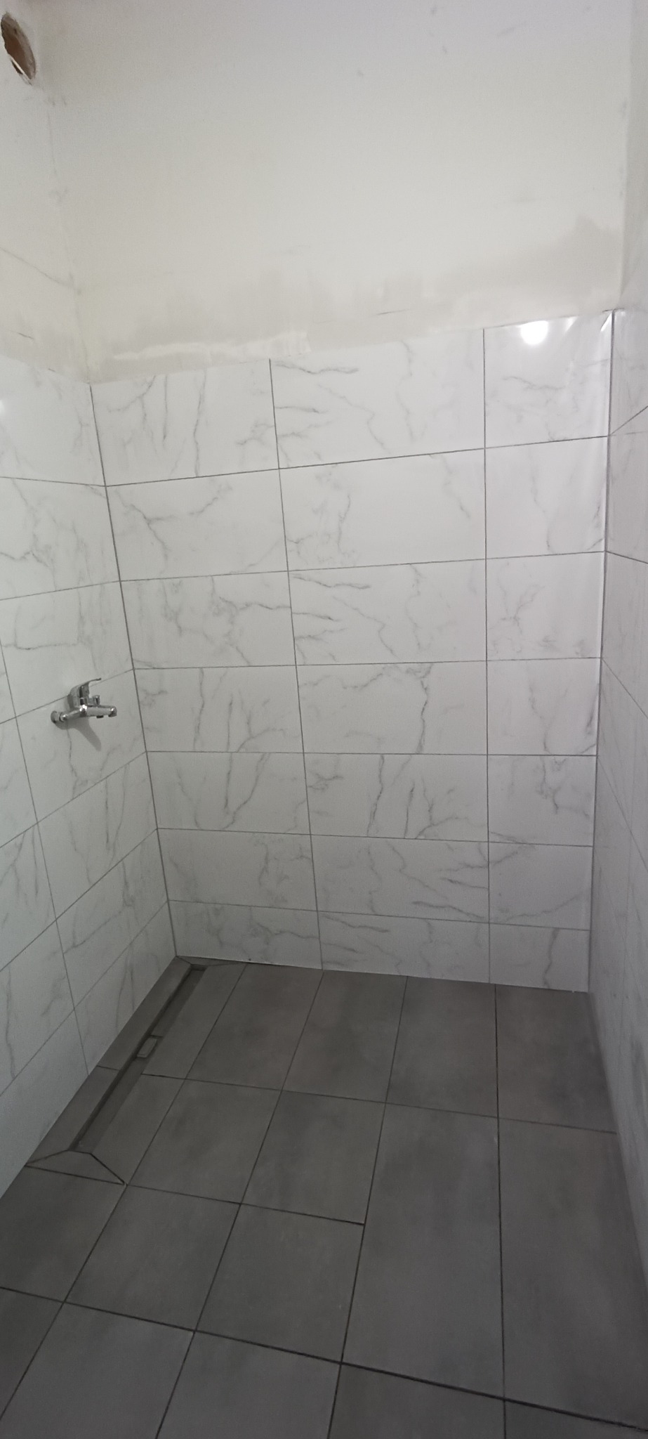 Wnętrze kabiny prysznicowej wyłożonej białymi płytkami imitującymi marmur na ścianach i szarymi płytkami na podłodze z odpływem liniowym.