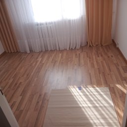 LukasBuilds - Jasny pokój z nowo ułożonymi panelami podłogowymi w odcieniach brązu, widoczne zasłony w oknie i fragment dywanu z niebieską taśmą zabezpieczającą.