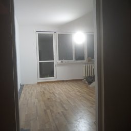 Remonty Wnętrz Łukasz Wysocki - Widok z korytarza na jasne, odnowione wnętrze z drewnianą podłogą, białymi ścianami i trzema oknami, z widocznym grzejnikiem i elementami po remoncie.