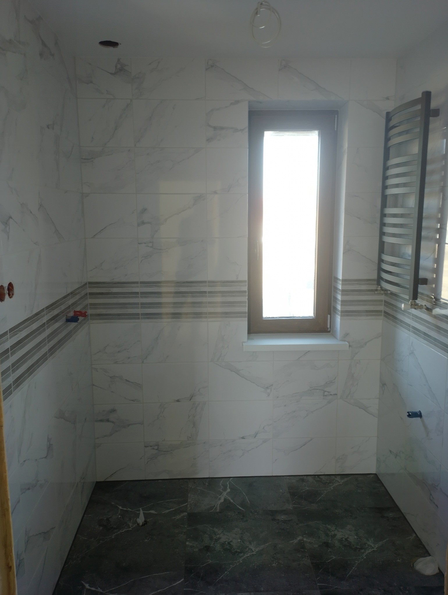 Wykończona łazienka z białymi płytkami imitującymi marmur na ścianach, pasem dekoracyjnym z szarymi paskami i ciemną podłogą z płytek. Grzejnik drabinkowy w rogu, okno z jasnym światłem w tle.