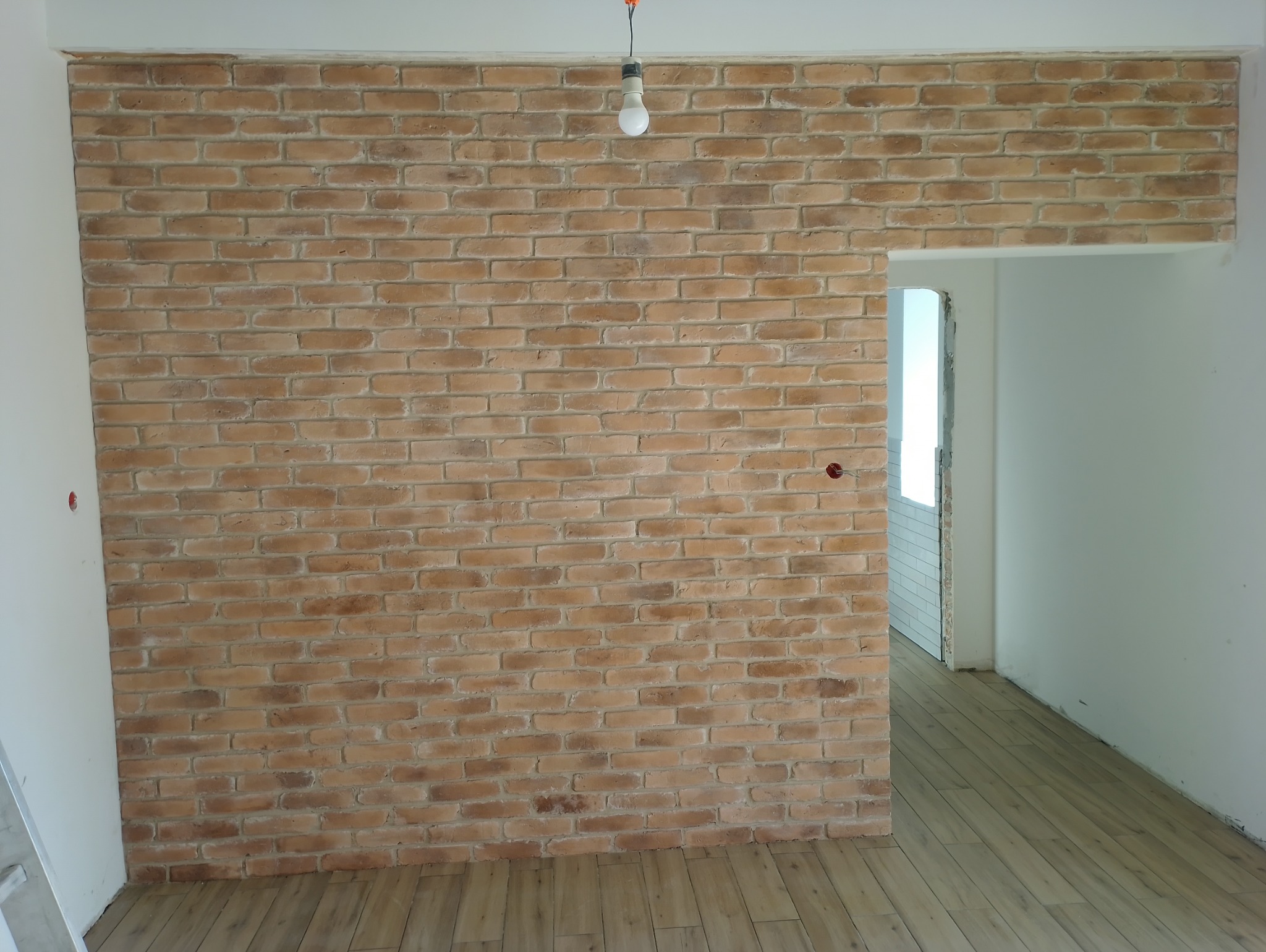 Ściana wyłożona dekoracyjną cegłą w odcieniach beżu i brązu, widoczne gniazdka elektryczne, surowa żarówka na kablu i fragment drewnopodobnej podłogi w trakcie prac wykończeniowych.