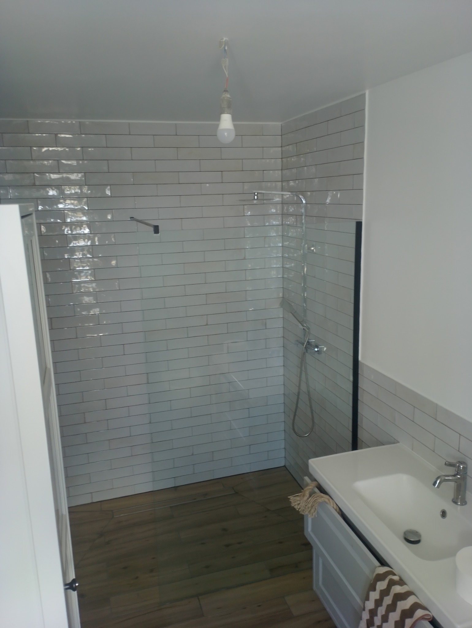 Nowoczesna łazienka z białymi, błyszczącymi płytkami ułożonymi w cegiełkę, kabiną prysznicową ze szklaną ścianką, drewnopodobną podłogą i białą umywalką z szarą szafką, z wiszącą żarówką bez klosza.