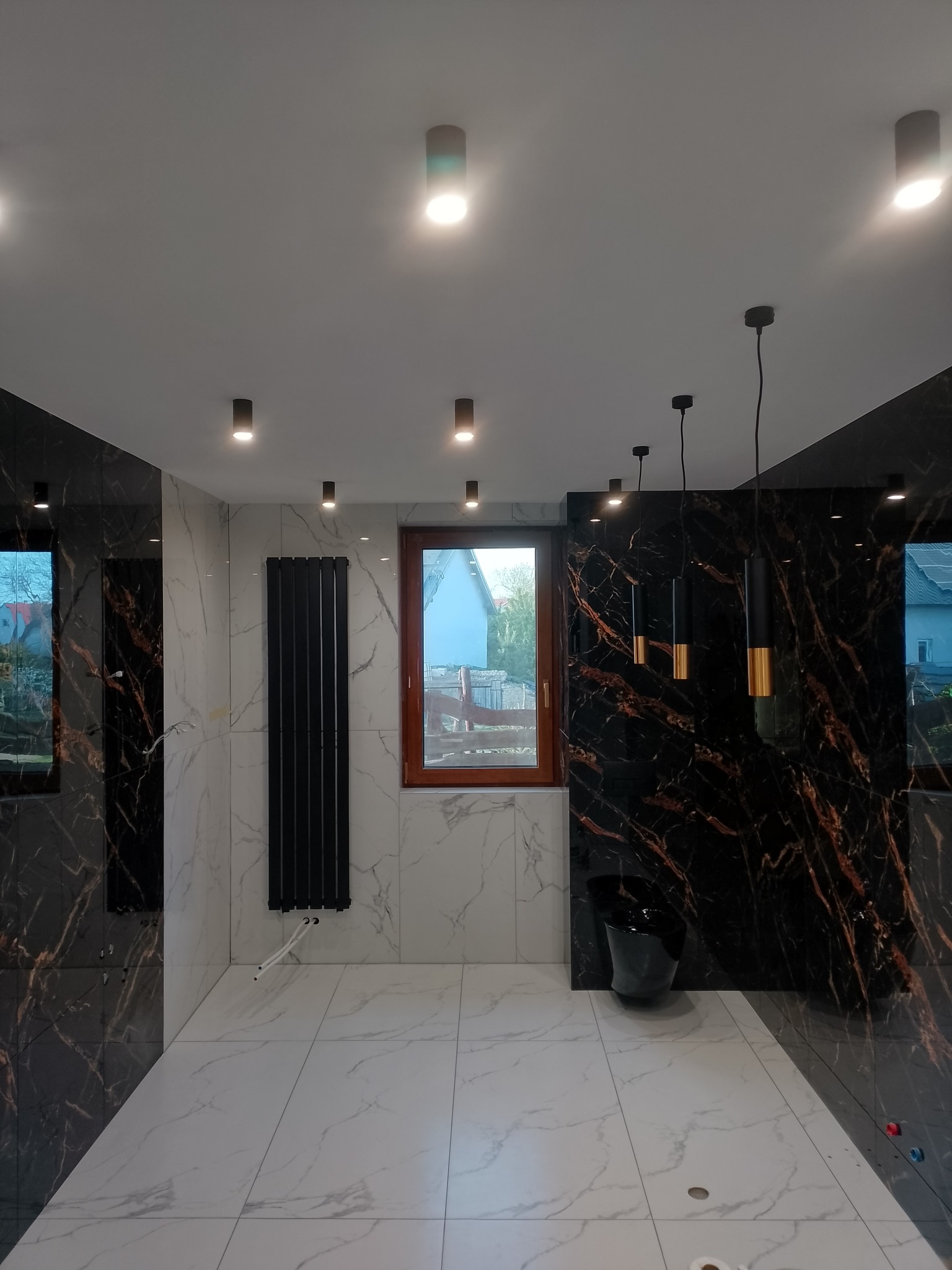 Elegancka łazienka z marmurowymi płytkami, czarnym grzejnikiem i wiszącą toaletą. Nowoczesne oświetlenie punktowe i designerskie lampy wiszące.