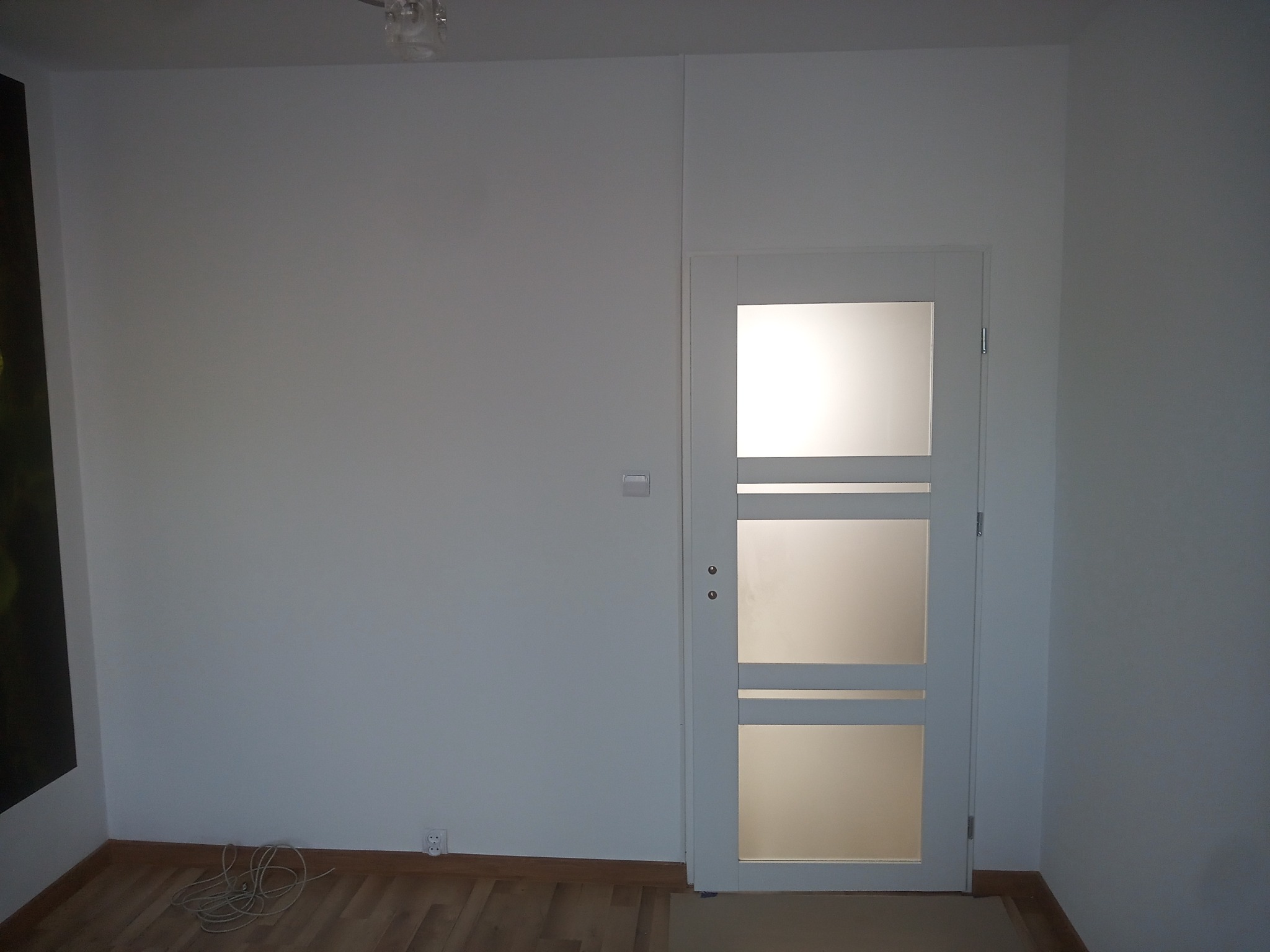 Białe drzwi z trzema przeszkleniami w nowo wykończonym, jasnym pokoju z drewnianą podłogą i białymi ścianami; widoczny zwisający kabel elektryczny.