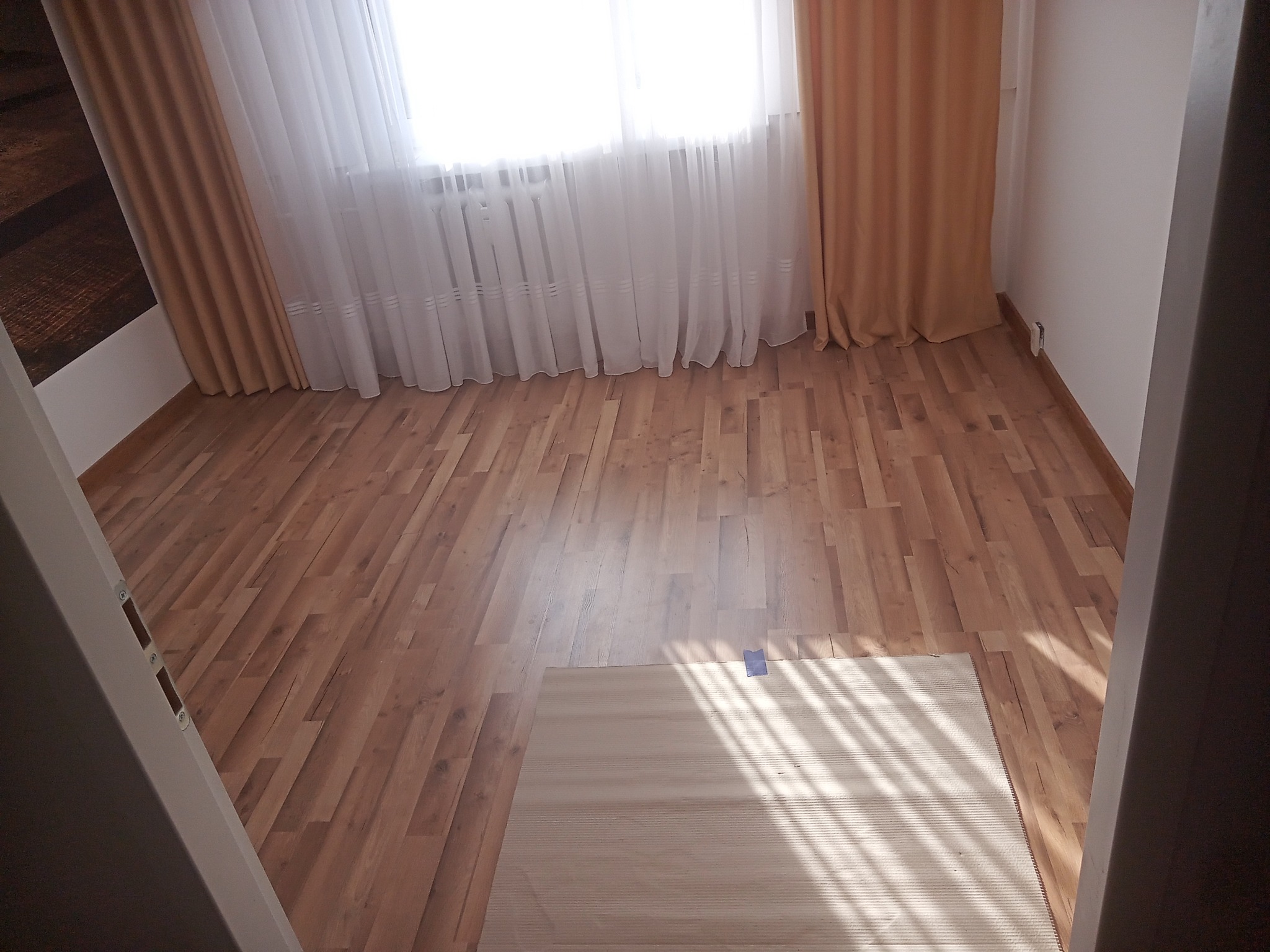 Jasny pokój z nowo ułożonymi panelami podłogowymi w odcieniach brązu, widoczne zasłony w oknie i fragment dywanu z niebieską taśmą zabezpieczającą.