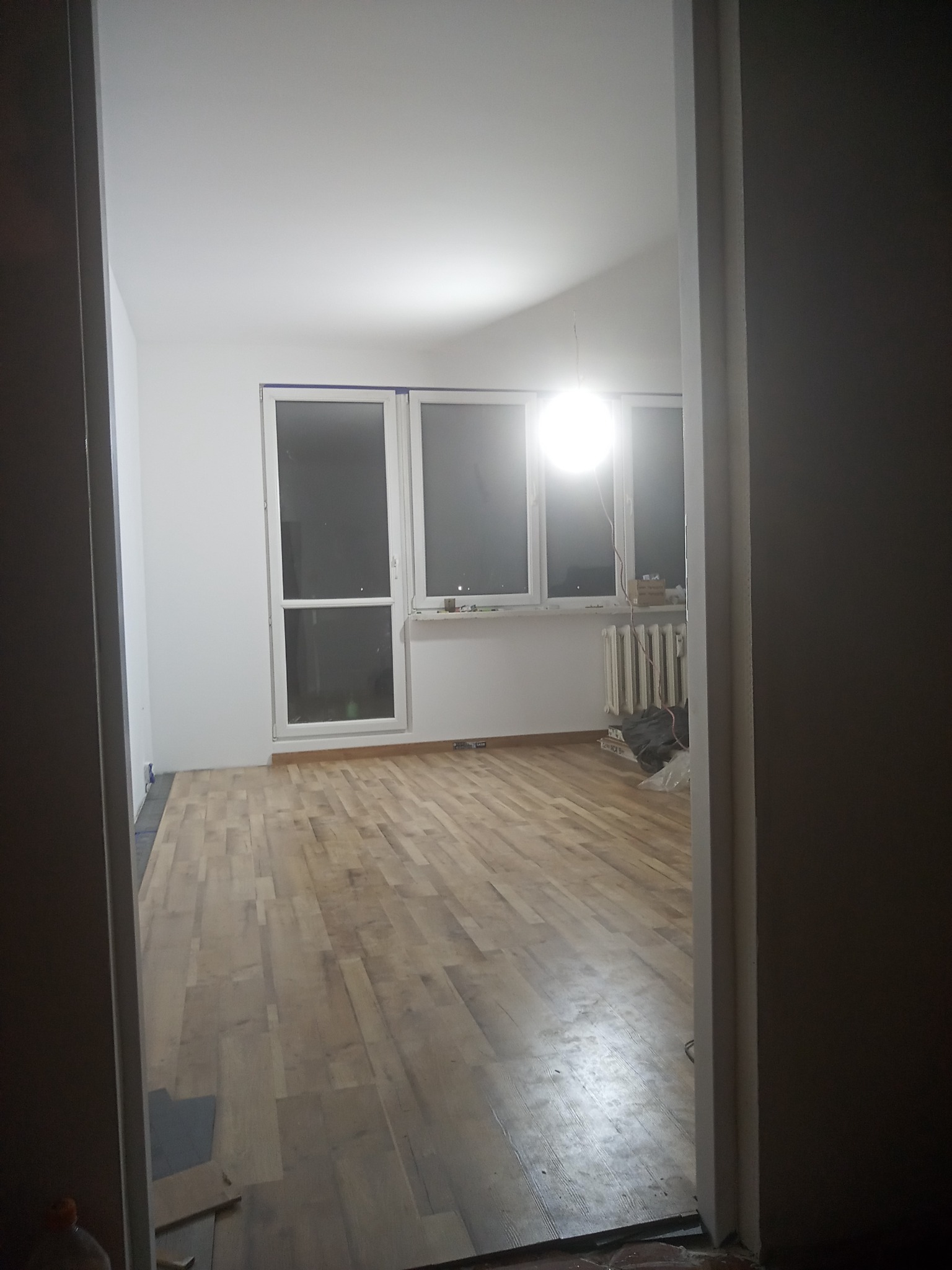 Widok z korytarza na jasne, odnowione wnętrze z drewnianą podłogą, białymi ścianami i trzema oknami, z widocznym grzejnikiem i elementami po remoncie.