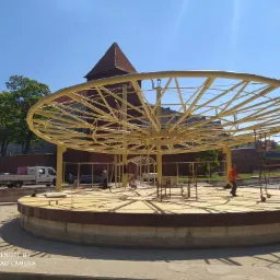 Montaż ażurowej, okrągłej konstrukcji z żółtych rur na betonowym podeście; w tle ceglana wieża i brygada pracowników w kamizelkach odblaskowych.