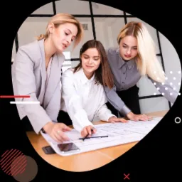 Trzy kobiety pochylają się nad projektem architektonicznym na drewnianym stole, używając ołówka i smartfona, w tle przeszklona ściana biurowa.