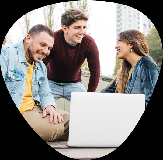 Trzy osoby uśmiechnięte, pochylone nad białym laptopem na zewnątrz, prawdopodobnie omawiające projekt.