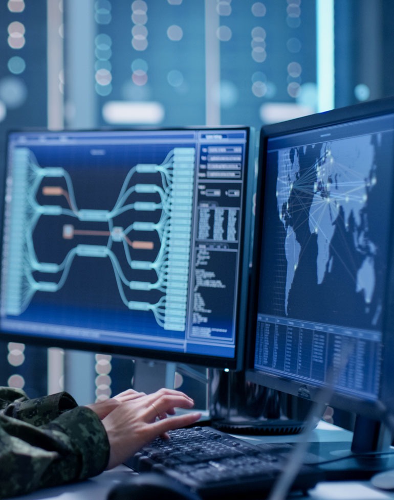 Osoba w mundurze wpisuje dane na klawiaturze komputera, na dwóch monitorach wyświetlane są schematy połączeń sieciowych i mapa świata z zaznaczonymi punktami połączeń.