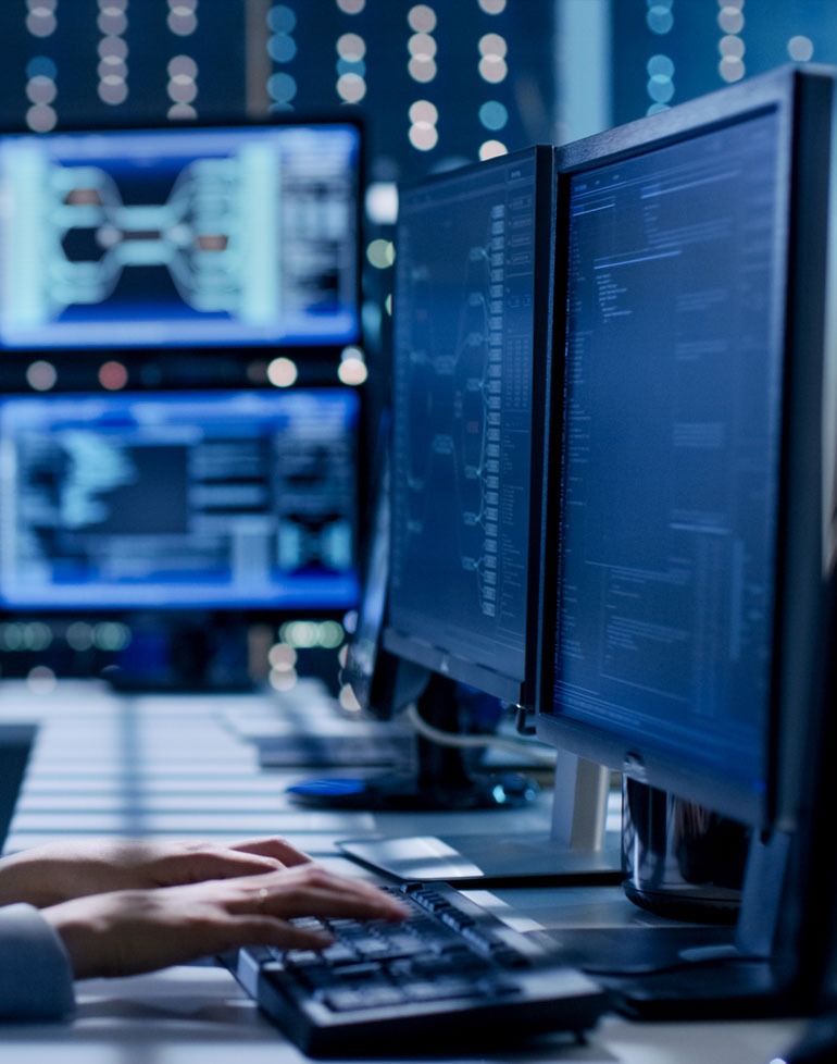 Programista pisze kod na dwóch monitorach w biurze, w tle widać dodatkowe monitory z diagramami i kodem, oświetlenie biurowe w tle tworzy efekt bokeh.